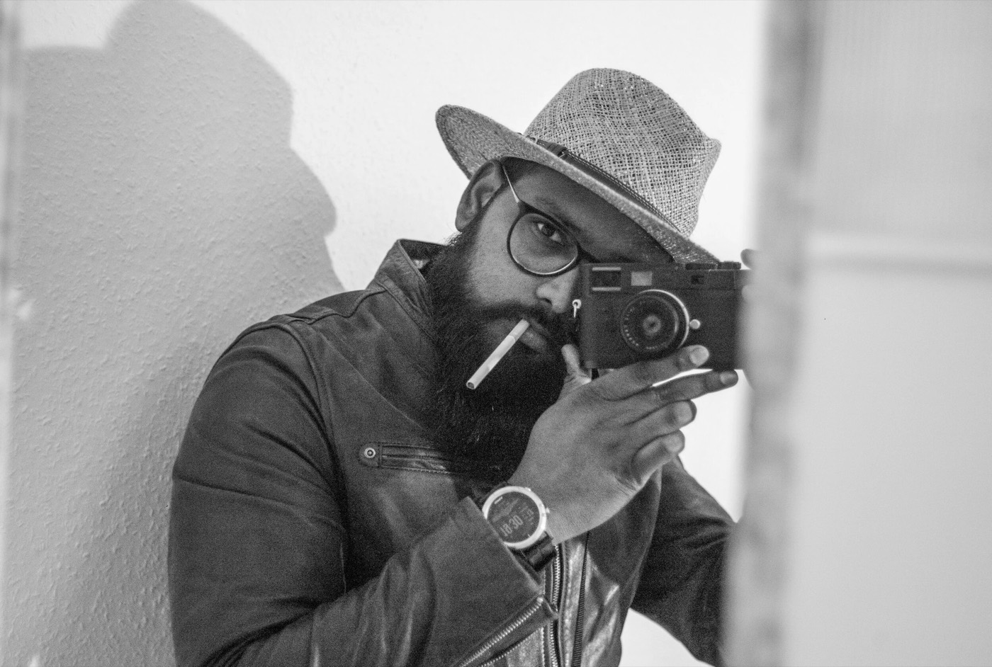 Bearded man in a straw trilby and glasses, cigarette in mouth, raising a rangefinder camera to his eye