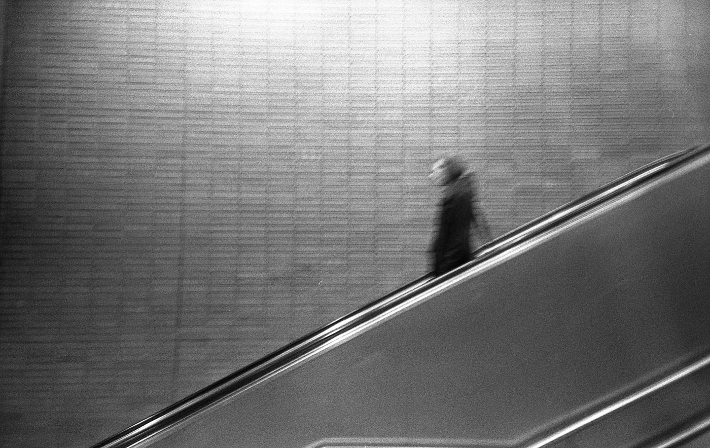 Lone blurred figure riding an escalator up past a vast tiled wall, diagonal handrail cutting the frame