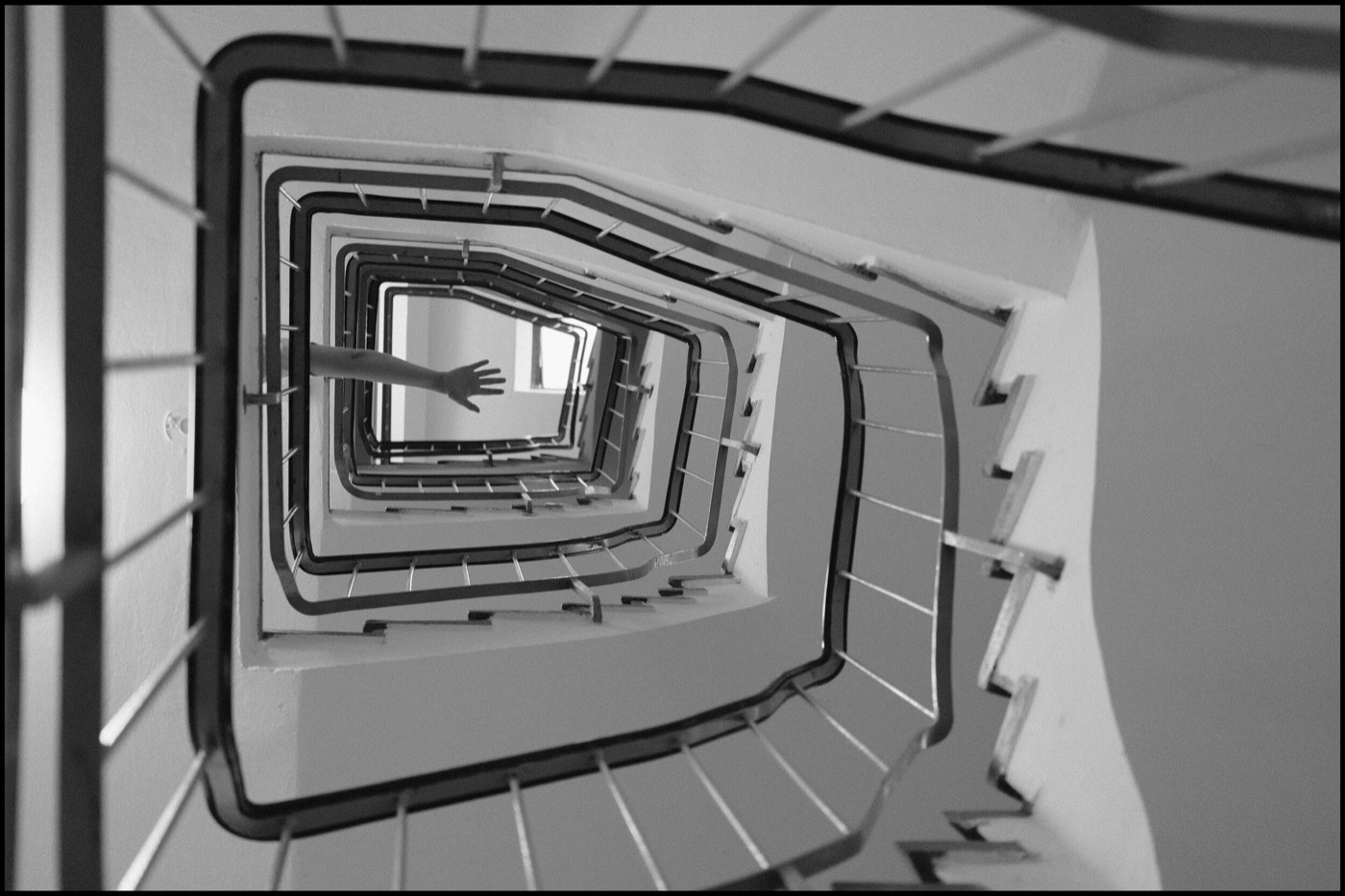 Looking up a rectangular spiral stairwell as a bare arm reaches out over the void several floors above