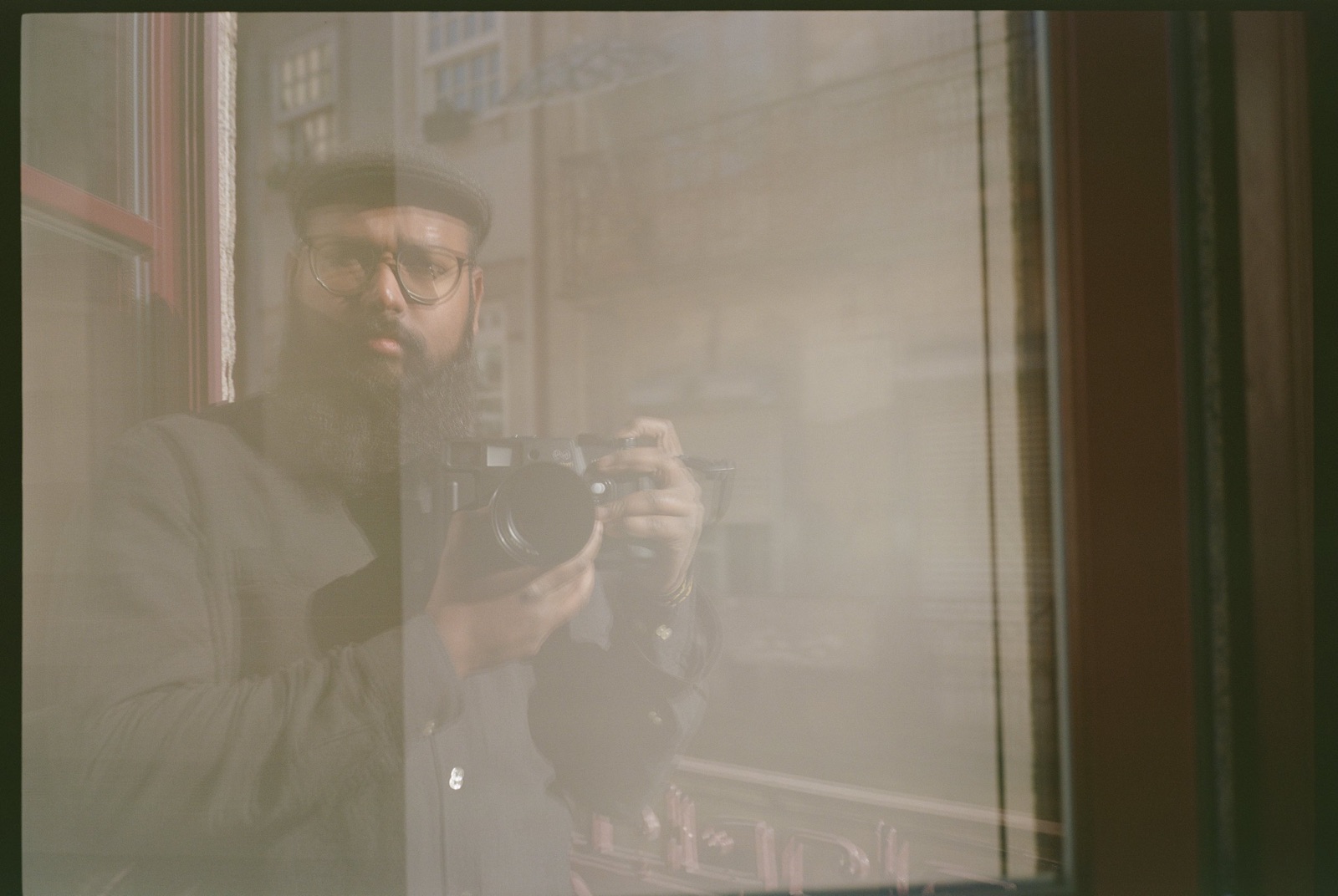 Bearded man in a flat cap and glasses reflected in a shop window, camera held at chest height