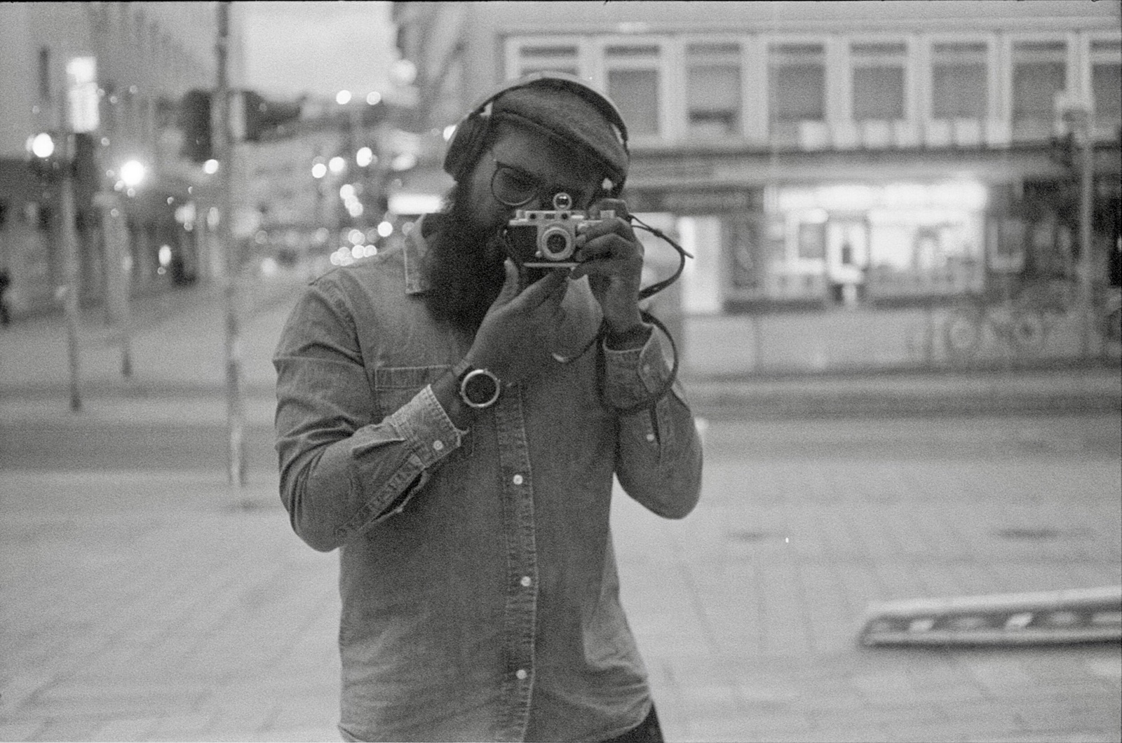 Bearded man in cap, glasses and denim shirt photographing his reflection on a city street at night