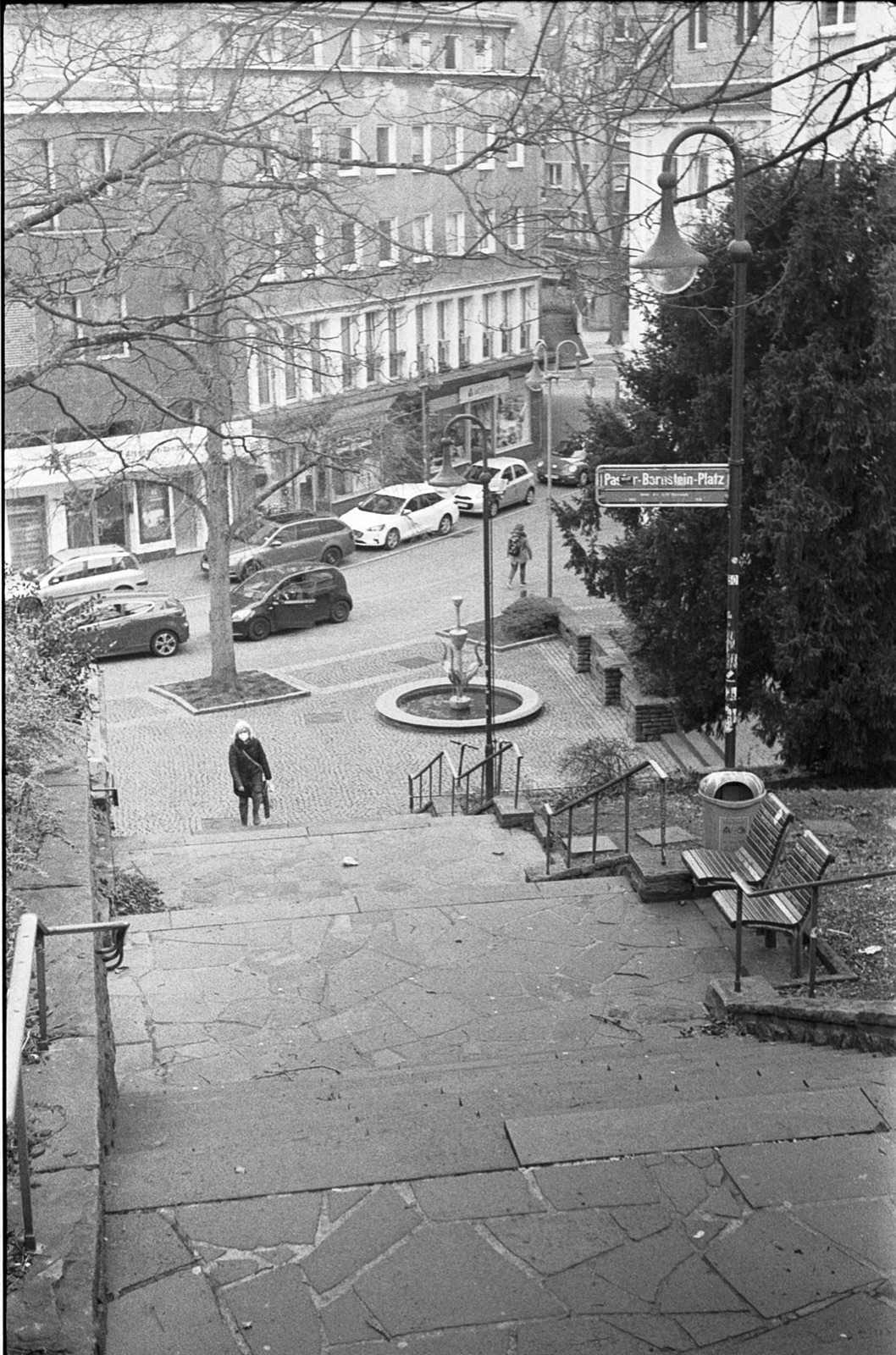 Looking down a flight of stone steps to a small square with a fountain, a lone woman crossing it in winter