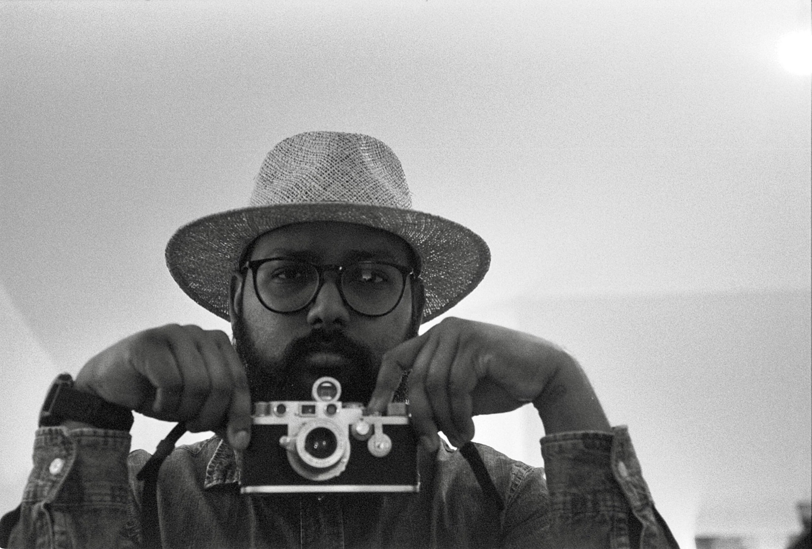 Bearded man in a straw hat and denim jacket holding a small camera at chest level, staring into the mirror