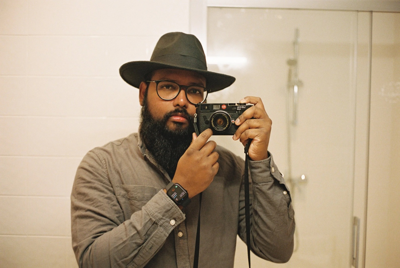 Bearded man in a wide-brimmed hat photographing himself in a bathroom mirror with a rangefinder