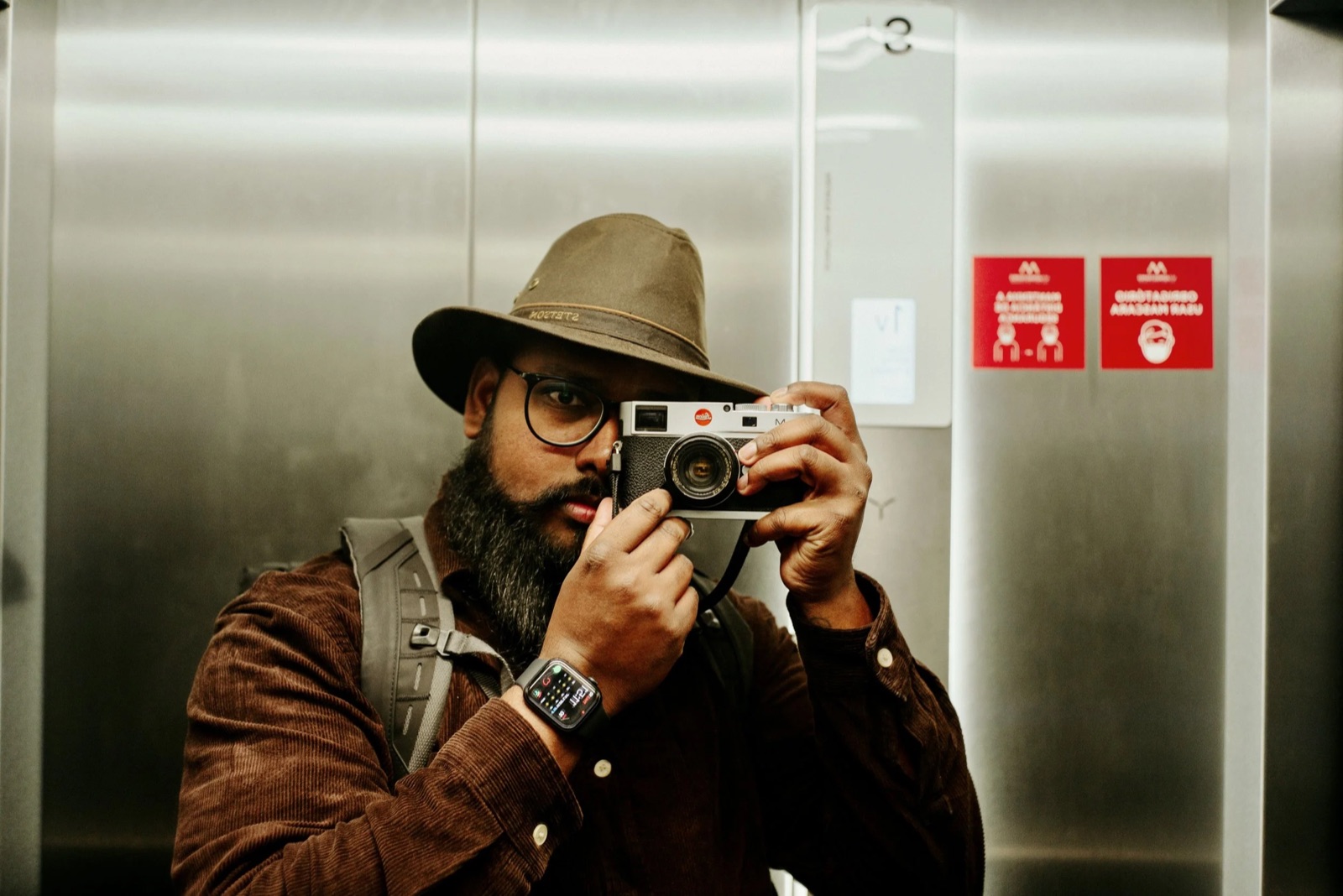 Bearded man in a wide-brimmed hat raising a camera to his eye, reflected in a steel lift wall