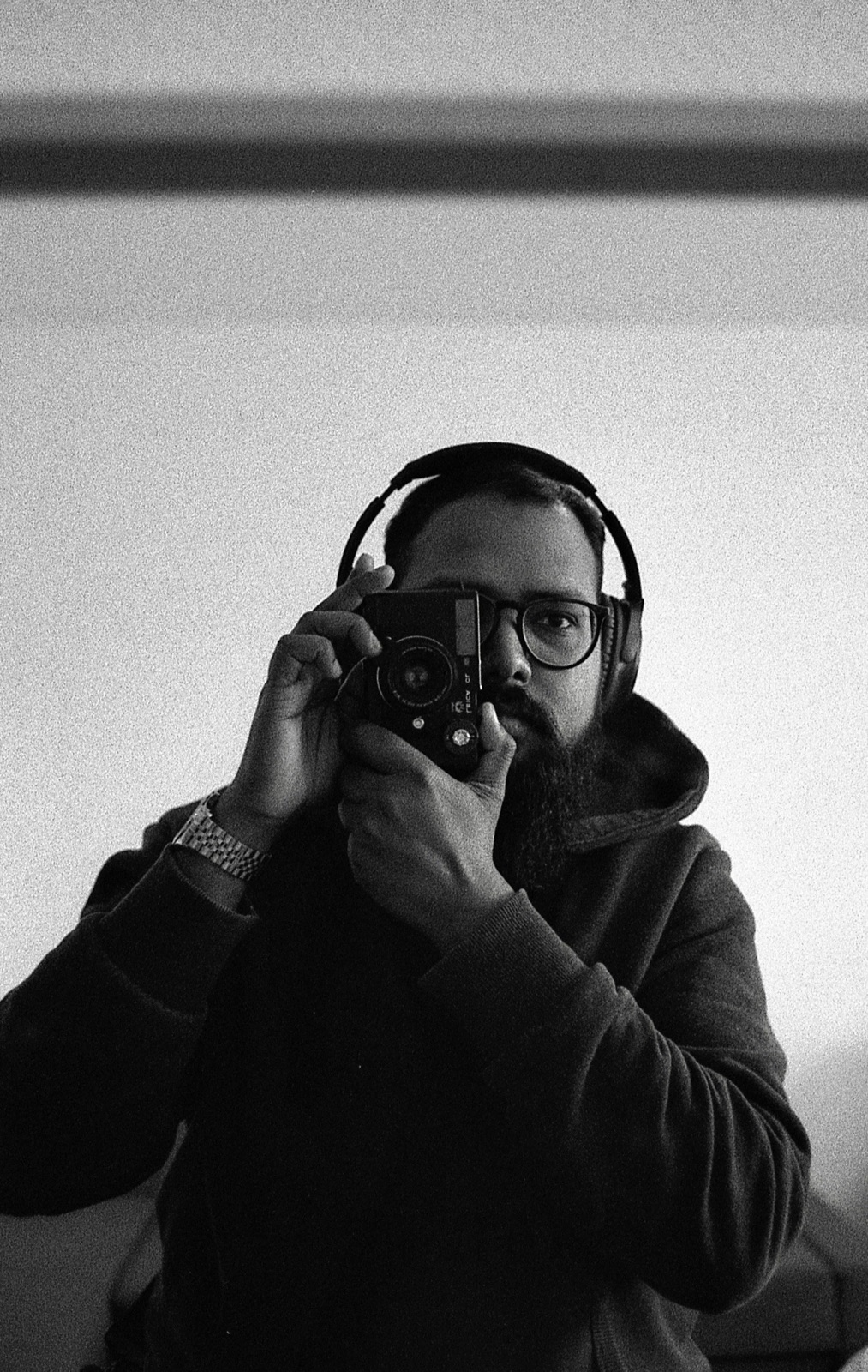 Grainy self-portrait of a bearded man in headphones raising a rangefinder camera to his eye