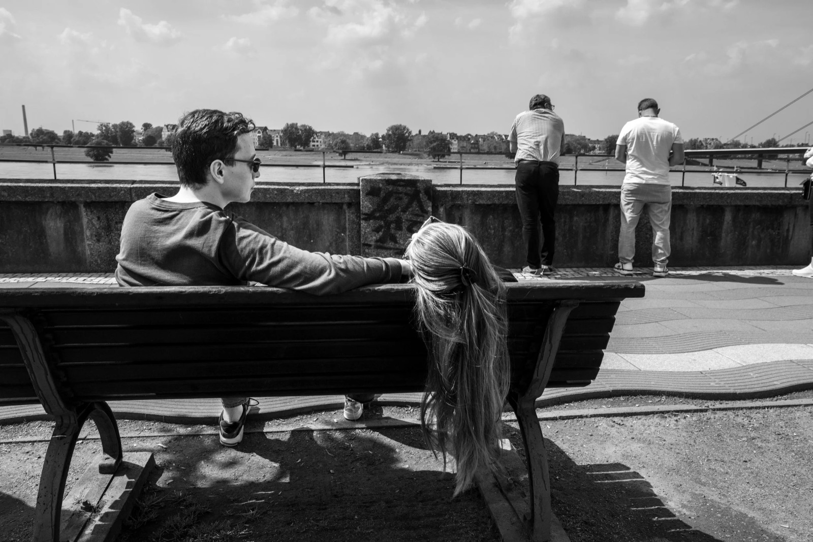 Man on a riverside bench with his arm around a woman whose long ponytail hangs over the backrest