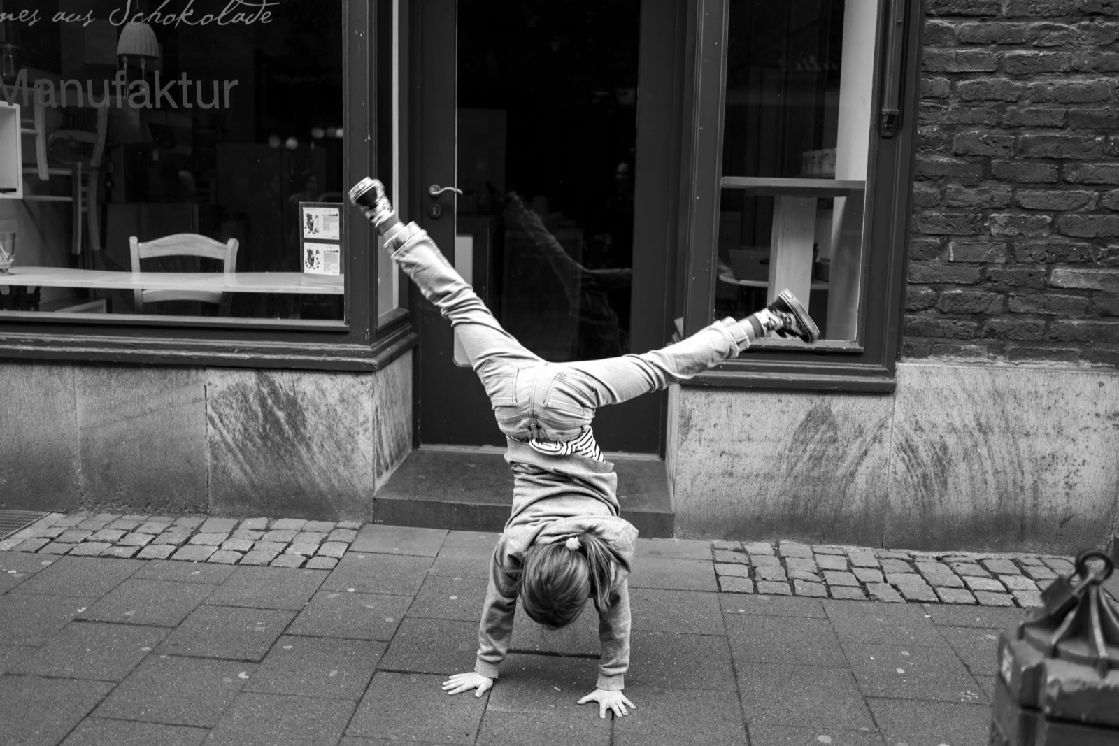 Child mid-cartwheel on cobblestones outside a shop window lettered Manufaktur