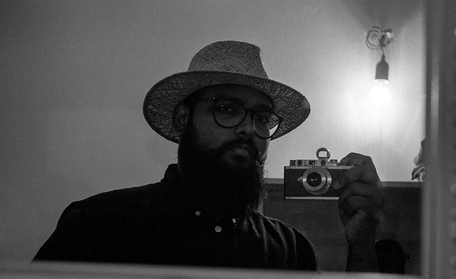 Bearded man in a straw hat and glasses holding an old rangefinder, bare bulb glowing behind him