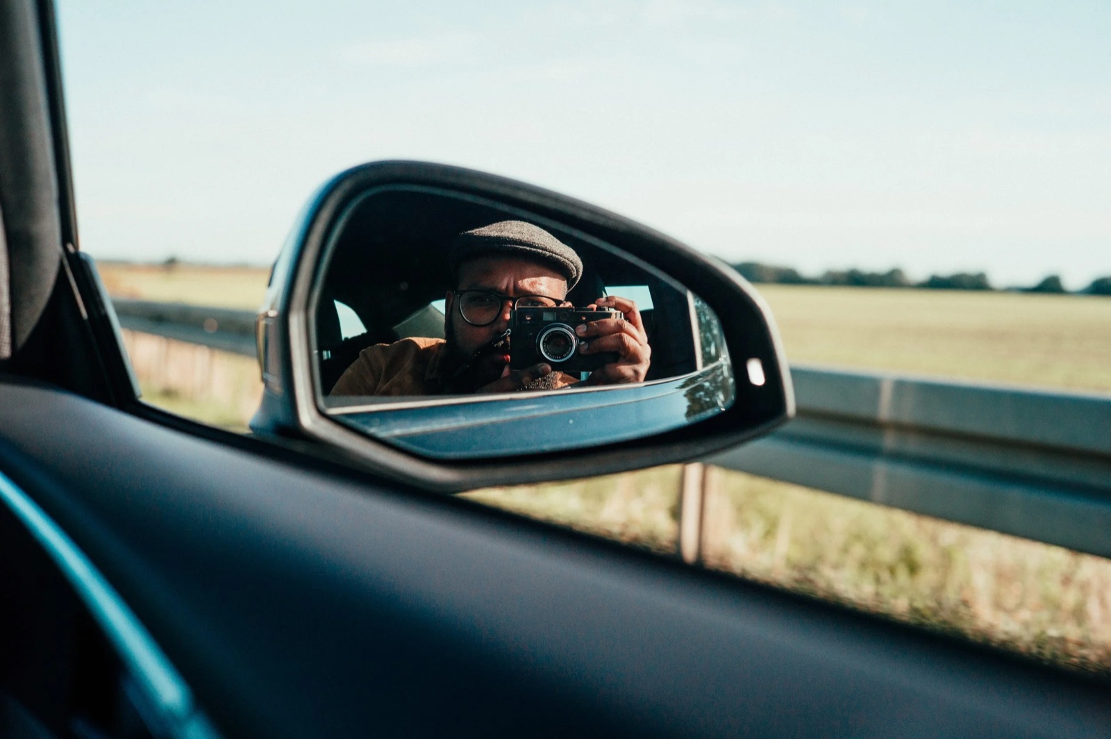 Man in a flat cap and glasses framed in a car wing mirror, fields and guardrail beyond