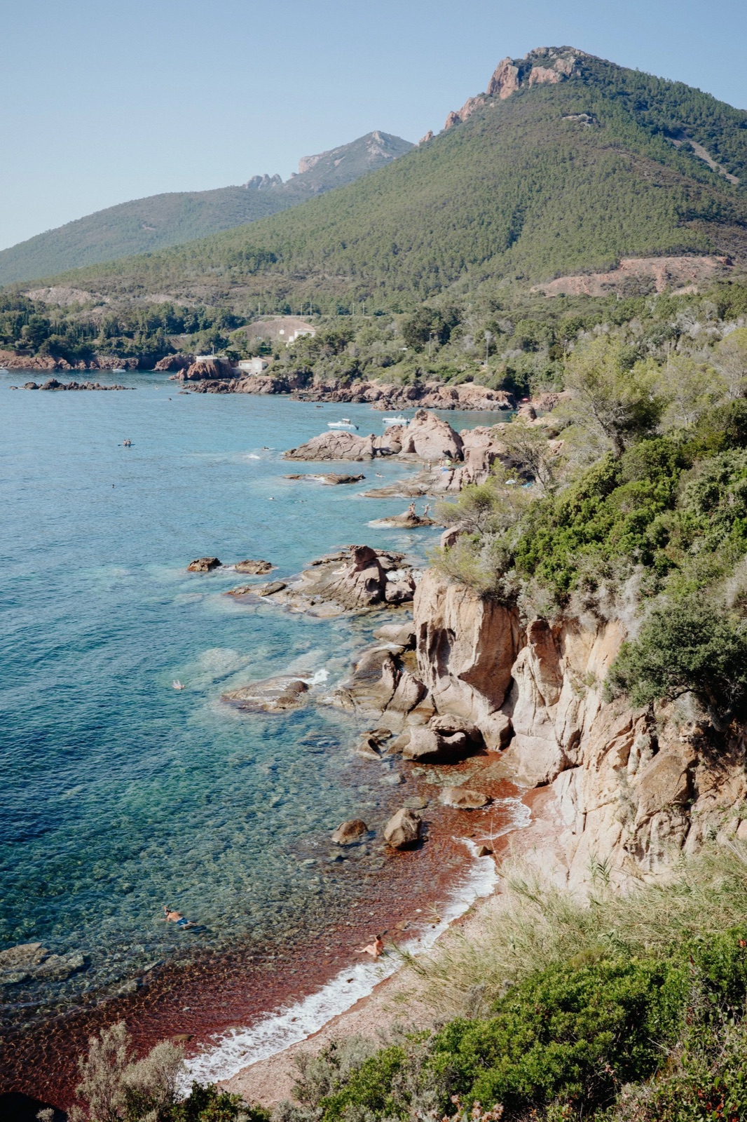 Red rocky coastline with turquoise shallows below a forested headland
