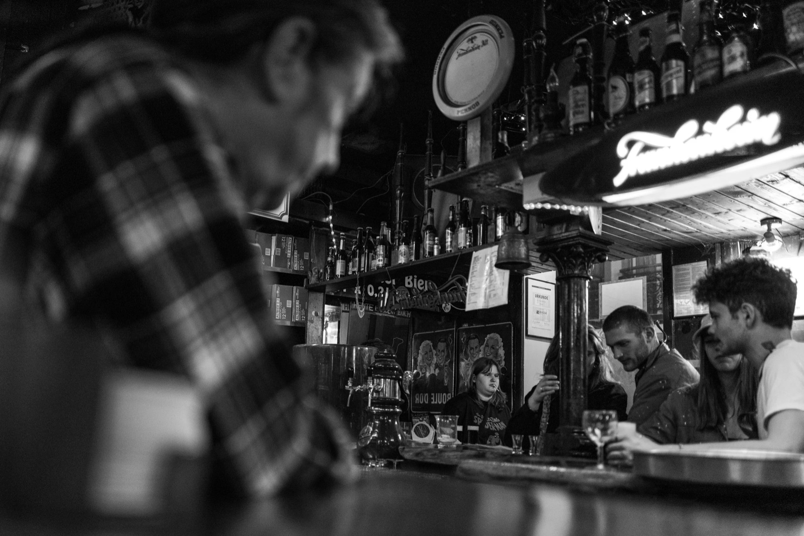 Bar interior with a blurred man in checked shirt in the foreground and drinkers under a neon beer sign