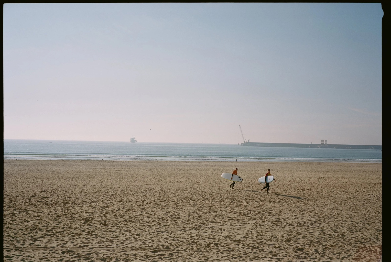 Two surfers in wetsuits carrying boards across a wide empty beach, cargo ship on the horizon