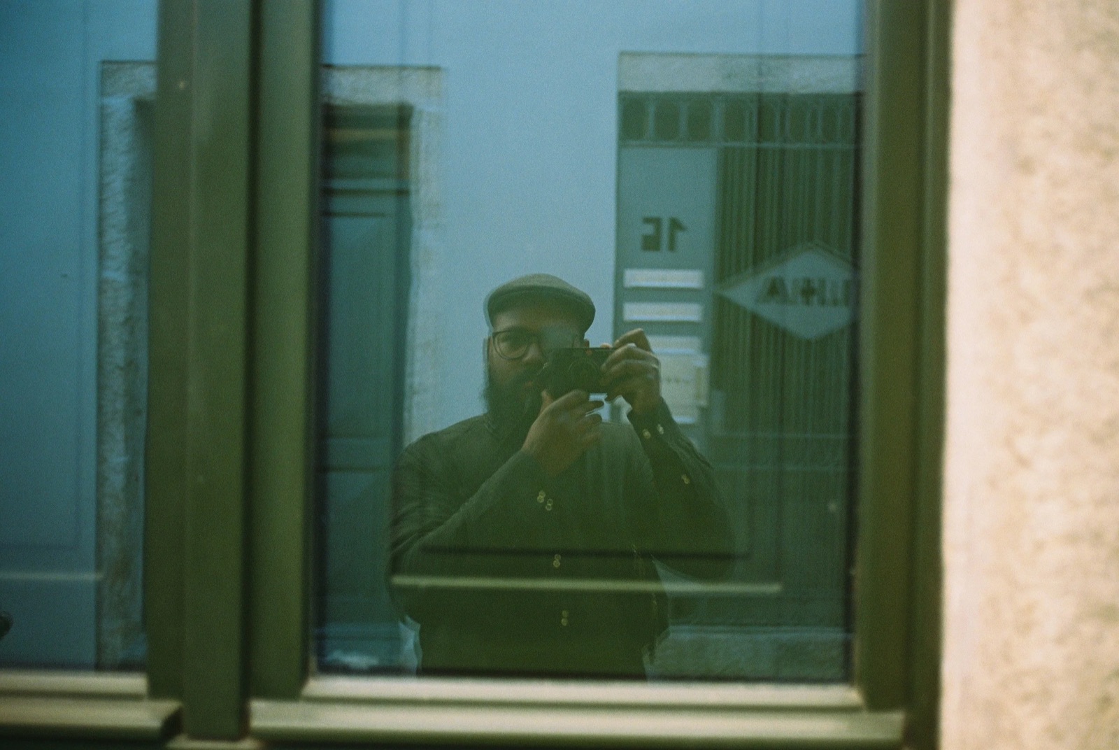 Photographer in a flat cap reflected in a window pane, camera at his eye, street doorway behind him