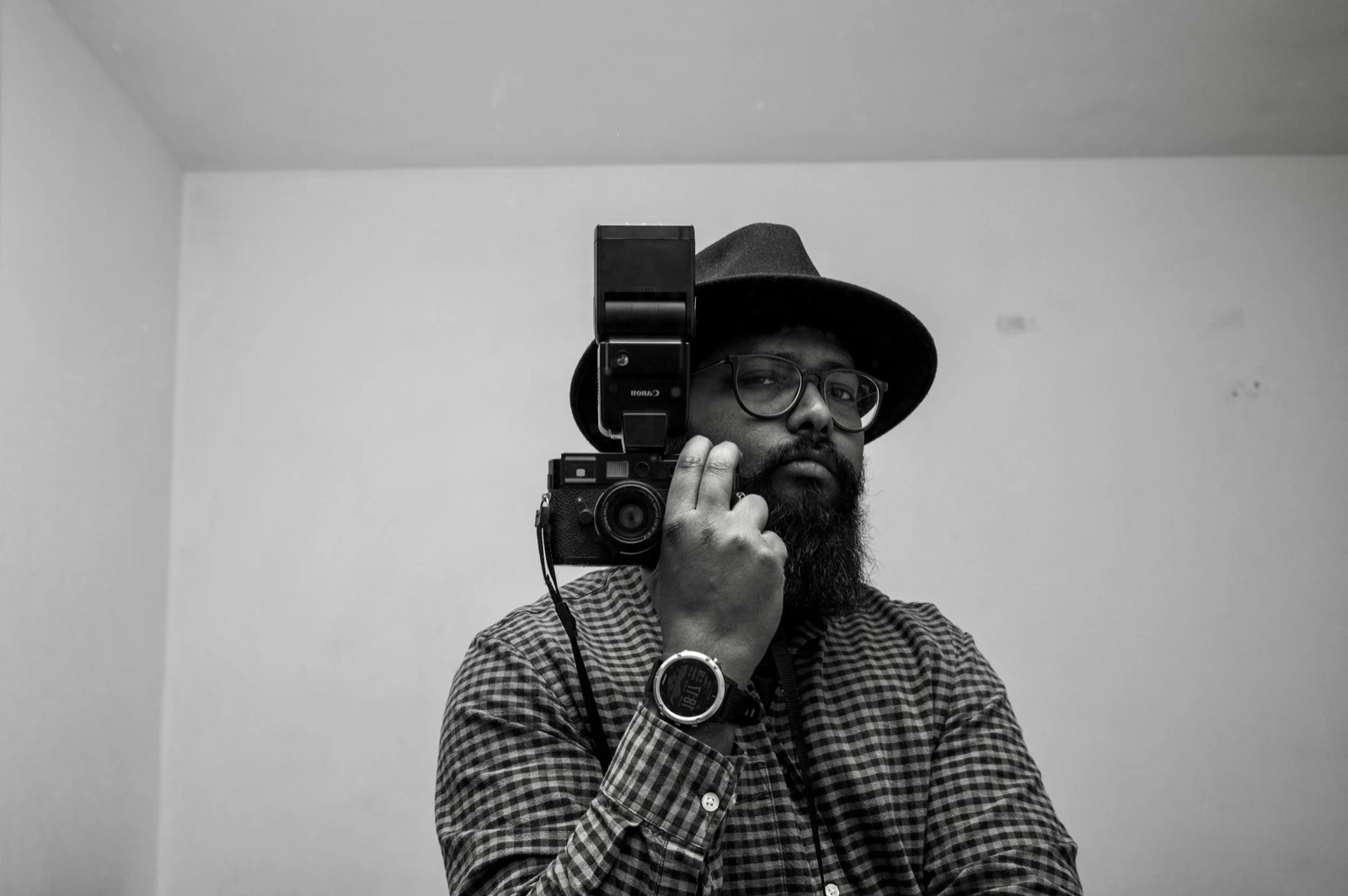 Bearded man in a hat and checked shirt holding a rangefinder with a flash beside his face