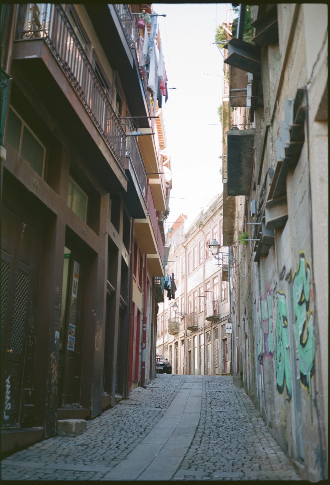 Narrow cobbled lane between tall tiled houses, laundry strung across balconies and graffiti on the right wall
