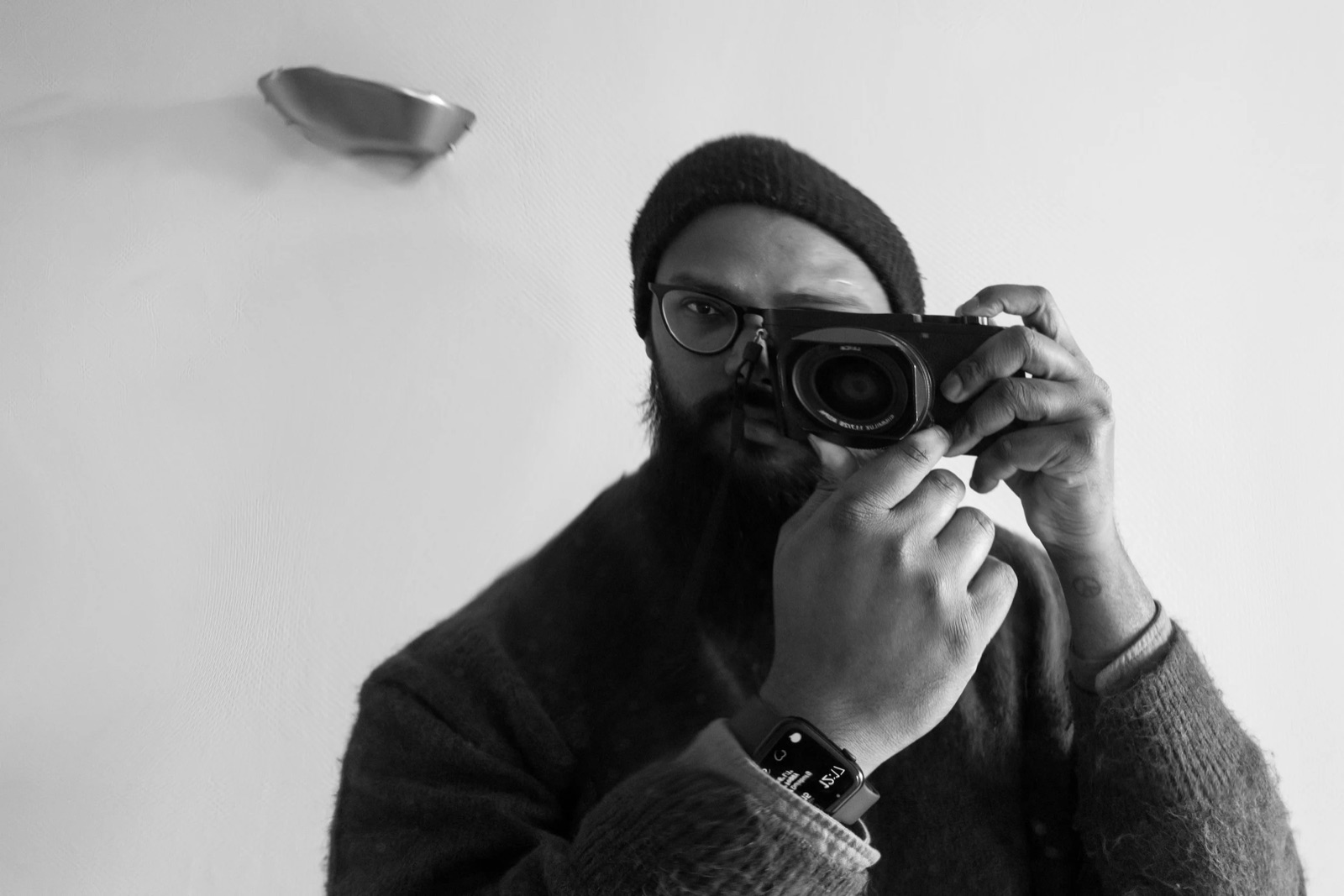 Bearded man in a beanie and knitted coat holding a compact camera to his eye against a white wall