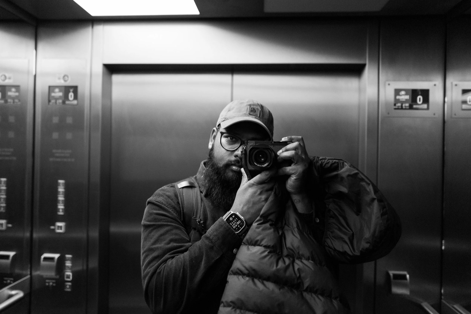 Bearded man in a cap and puffer vest photographing his reflection in a steel lift