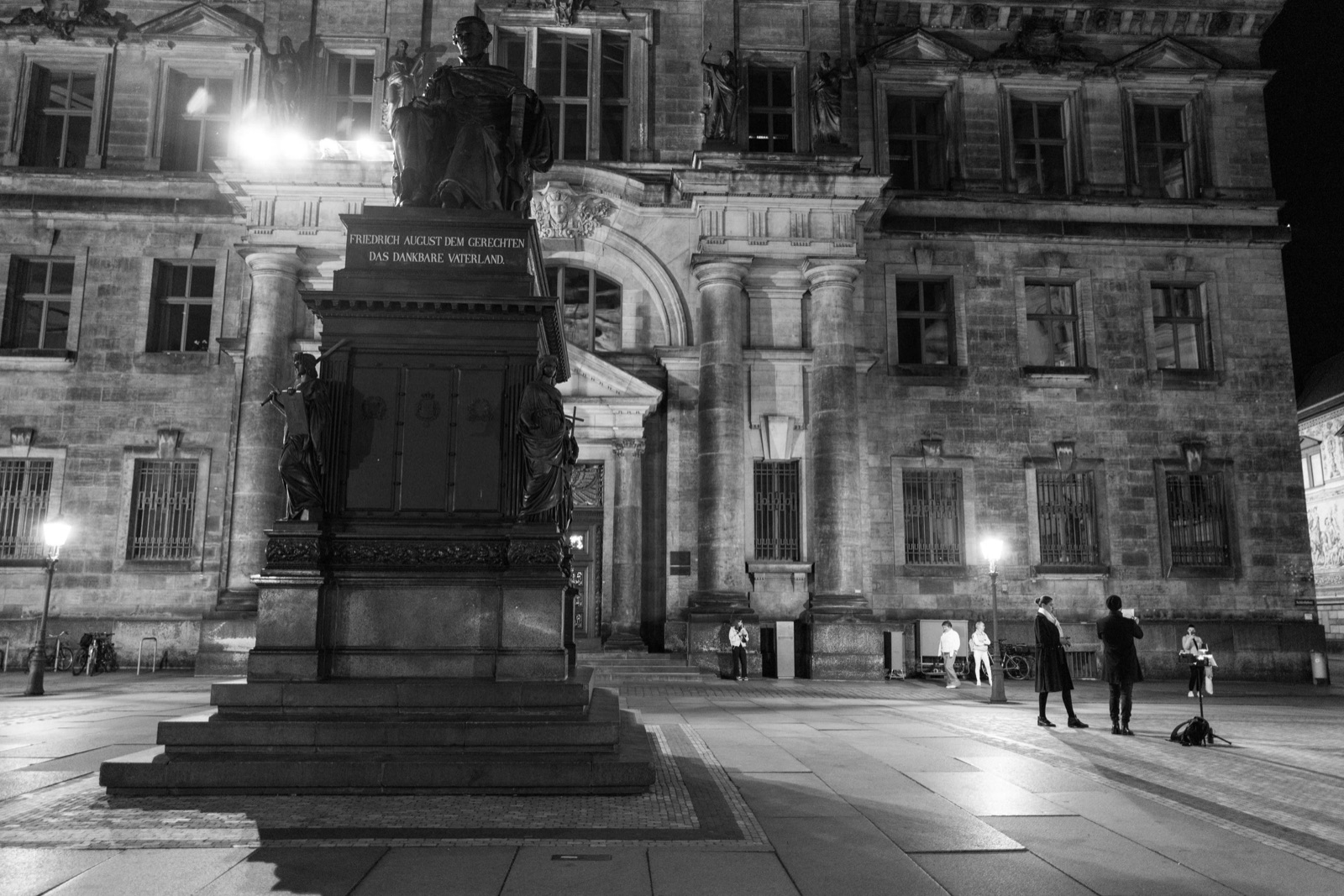 Bronze equestrian-style monument lit at night on an empty square before a floodlit baroque palace