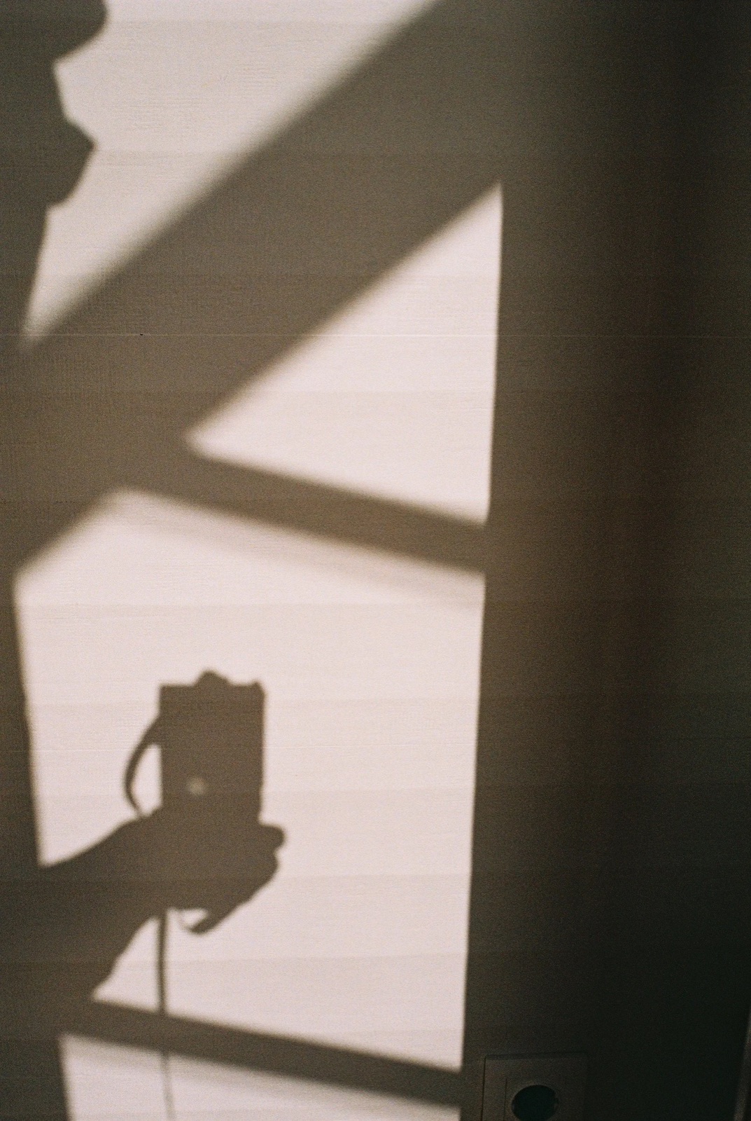 Shadow of hands holding a camera cast onto a wall beside a hard-edged window shadow