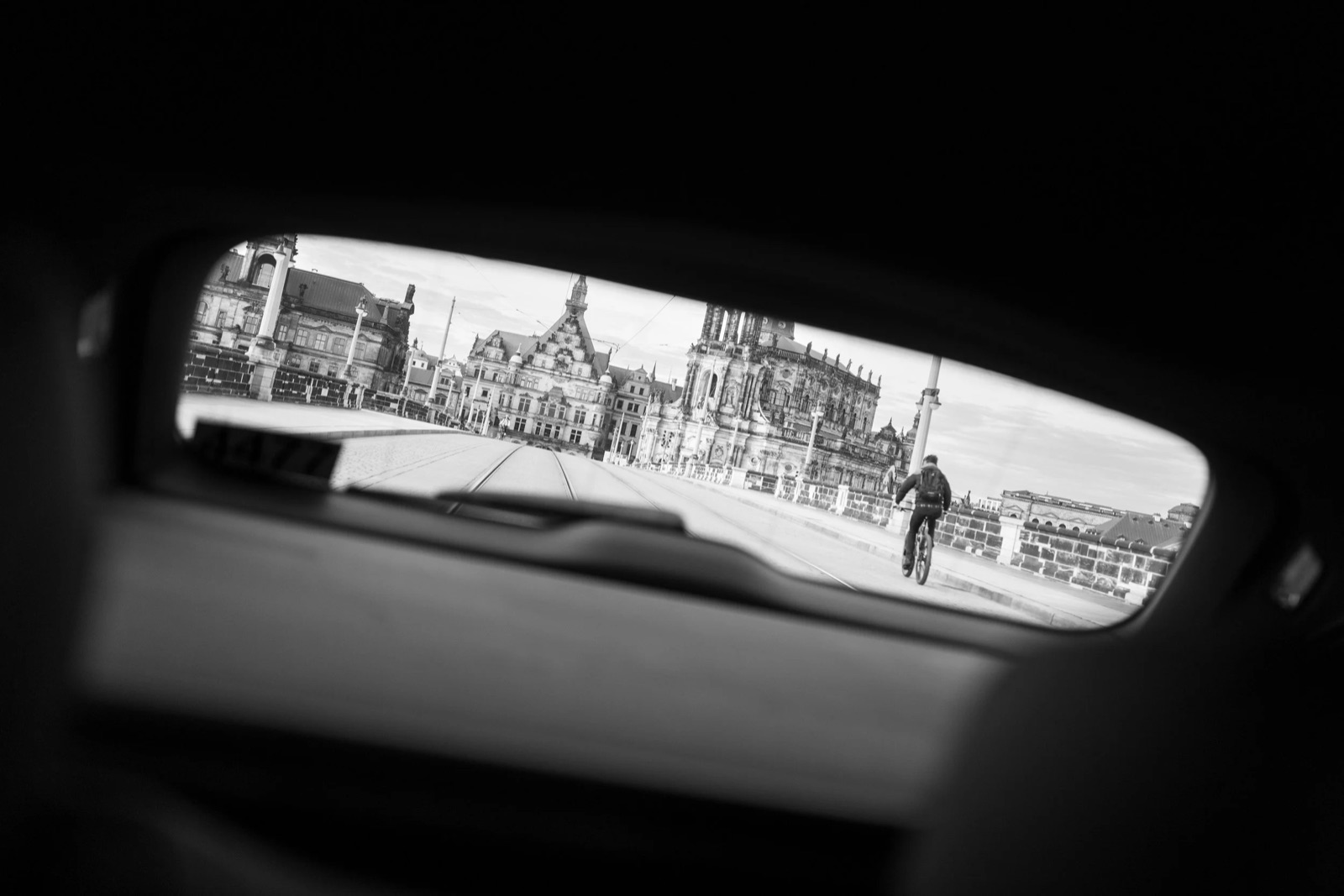Cyclist and baroque city skyline framed in the sliver of a car's side window, rest of frame black