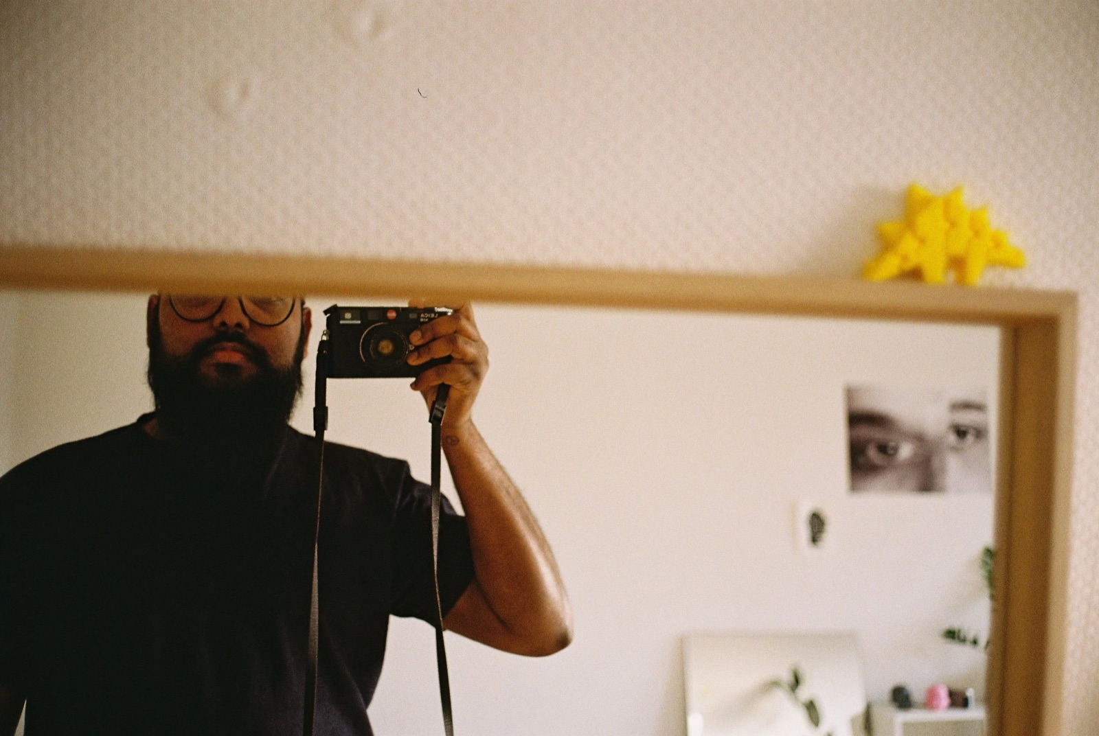 Mirror self-portrait of a bearded man in glasses raising a rangefinder camera to his eye in a bedroom