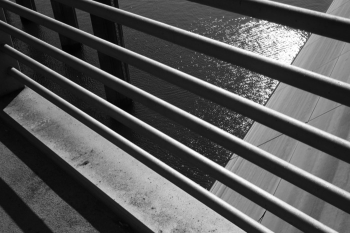 Diagonal metal railing bars slicing across sunlit rippling water and a concrete embankment
