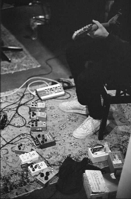 Guitarist's hands and white sneakers above a patterned rug strewn with effects pedals and cables