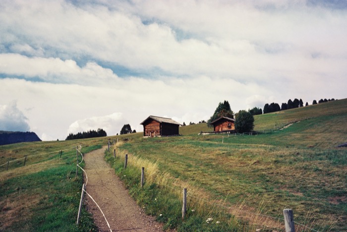 Fenced dirt path winding up an alpine meadow toward two dark wooden barns under piled cloud