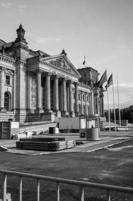 Columned parliament building behind construction barriers, stacked scaffolding and flagpoles in the foreground