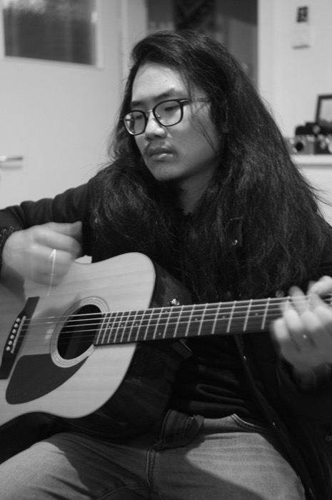 Long-haired man in glasses seated indoors, mid-strum on an acoustic guitar, gaze lowered
