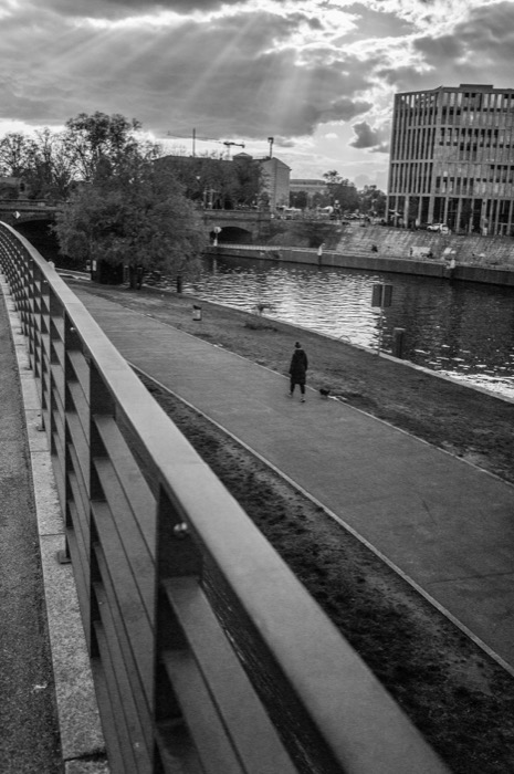 Person walking a small dog along an empty riverside promenade as sunbeams break through cloud