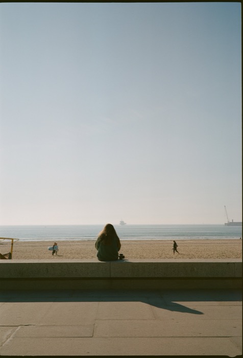 Woman sitting alone on a promenade wall above a wide empty beach, a ship far out on the horizon