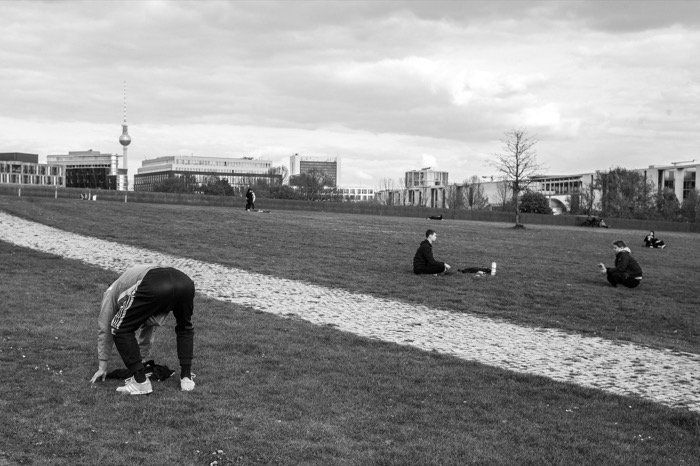 Person bending double to stretch on a park lawn while others sit spaced apart, TV tower on the skyline