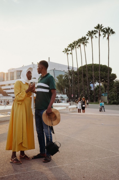 Couple talking on a sunlit seafront square, she in a long yellow dress, palms behind them