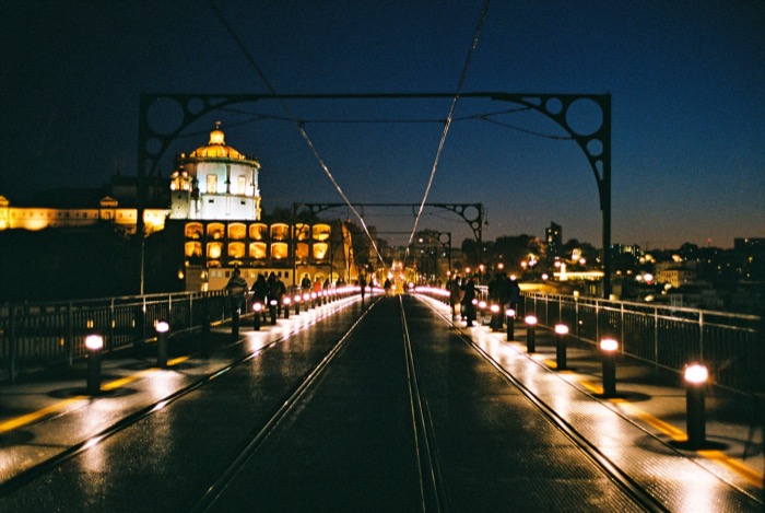 Tram tracks running across a lit iron bridge at dusk toward a domed church on the far bank