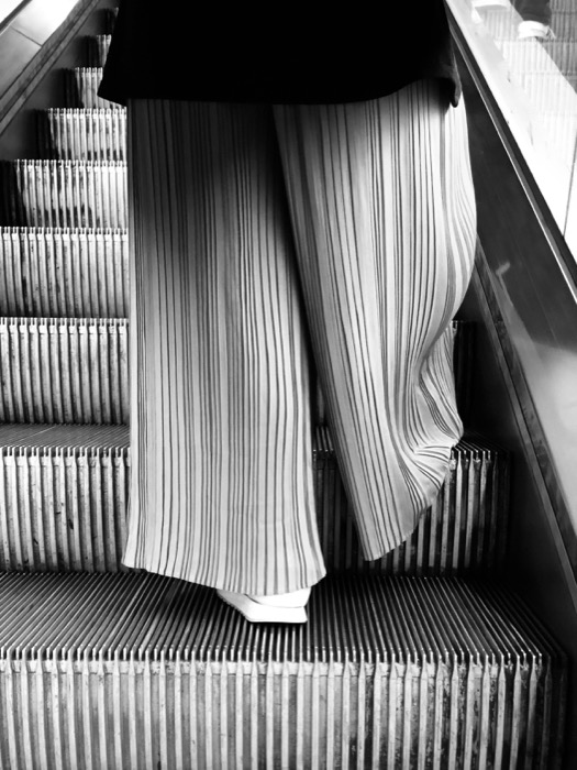 Pleated wide-leg trousers on an escalator, the fabric ribs echoing the metal step treads