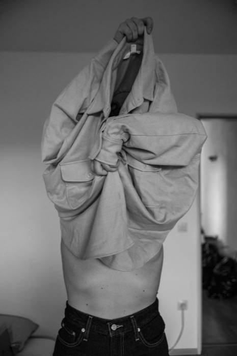 Bare-chested person pulling a shirt off over their head, face hidden by the fabric, in a bedroom