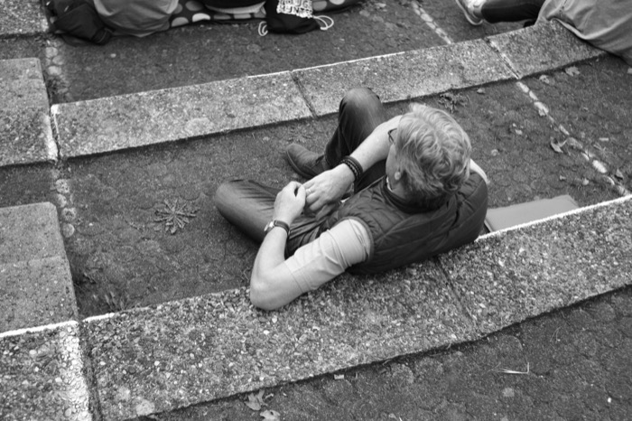Man with fair curly hair lying back on wide stone steps, seen from above, hands folded on his chest