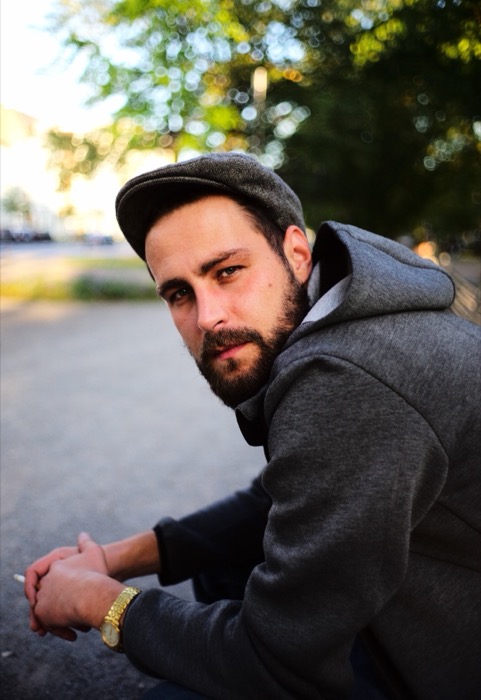 Bearded man in a flat cap and grey hoodie glances back over his shoulder, cigarette in hand