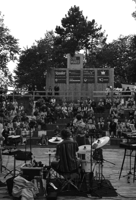 Drummer seen from behind on an open-air stage, playing to a packed hillside of seated spectators