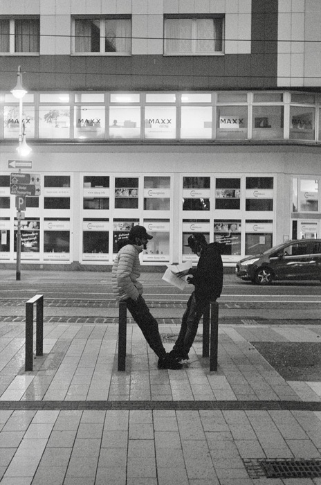 Two men leaning on bollards at night reading a newspaper together outside lit office windows