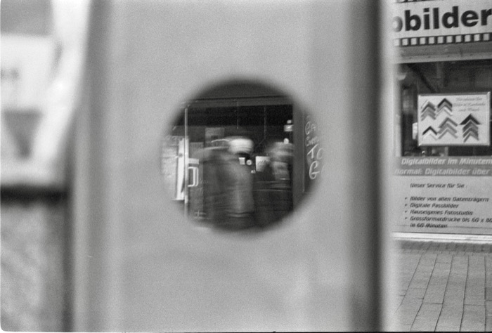 Blurred café scene glimpsed through a round hole in a street kiosk, photo-shop signage at the right
