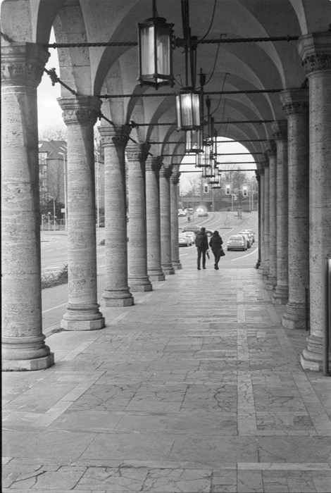 Couple walking away down a long stone colonnade with hanging lanterns, traffic beyond the arches