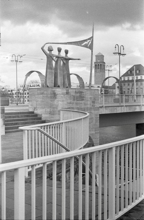 Bronze sculpture of three slender figures holding a flag on a plinth beside a bridge and white railings