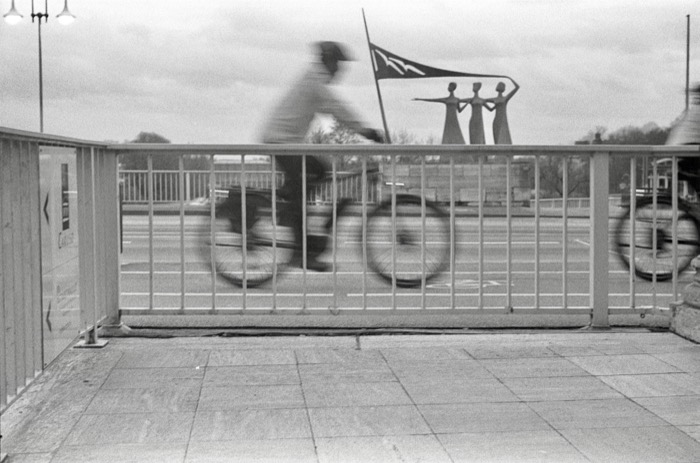 Cyclist blurred with motion crossing a bridge behind a railing, three tall figure sculptures beyond