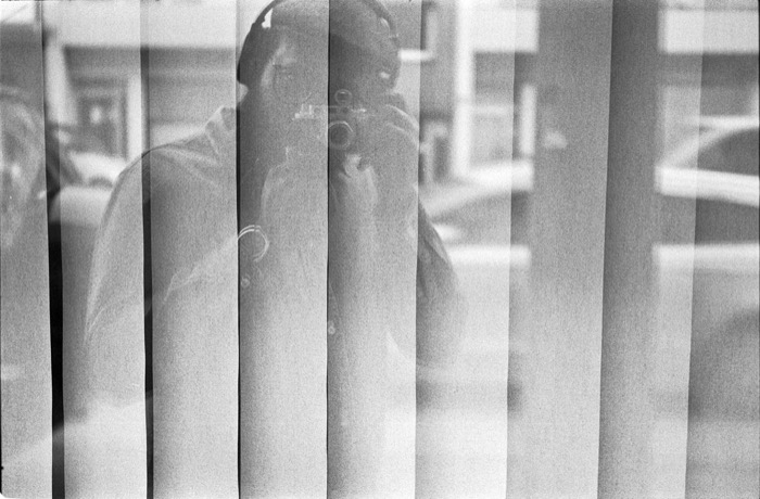 Reflection of a person aiming a camera, broken into vertical strips by window blinds