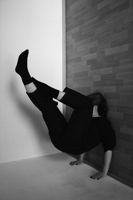 Woman in black balancing upside down on her hands in a bare room corner, legs against the wall