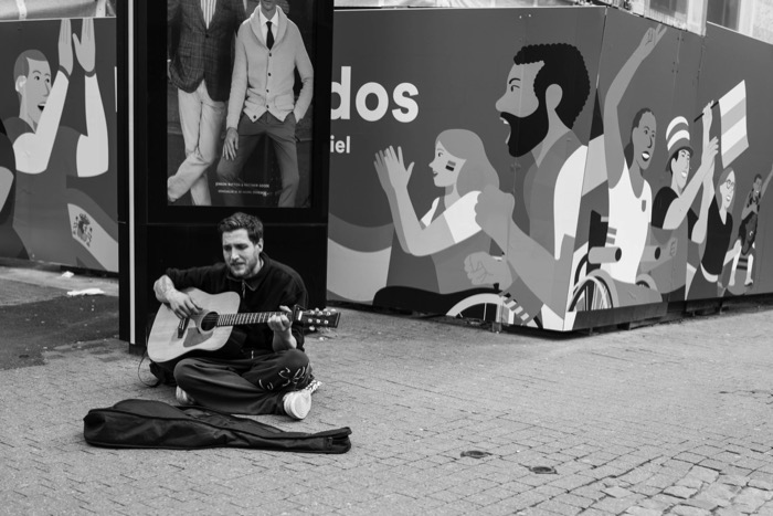 Busker sitting cross-legged with a guitar on paving, a mural of cheering figures behind him