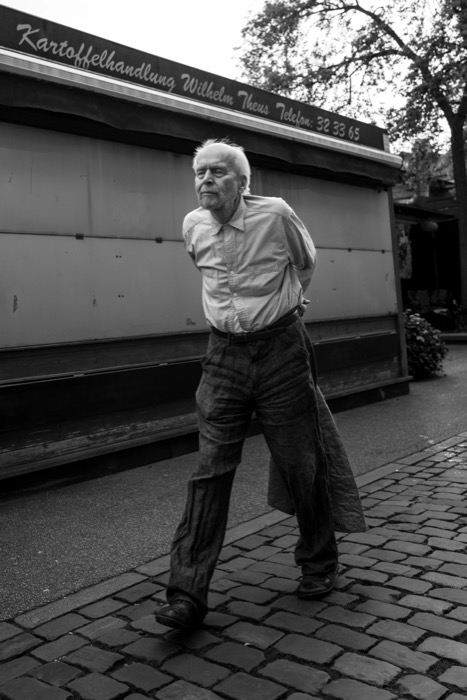 Elderly man in a striped shirt striding across cobblestones past a shuttered potato merchant's stall